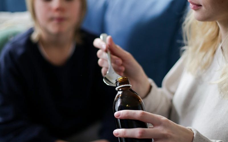 A caring mother pours medicine for her sick child sitting on a couch indoors.