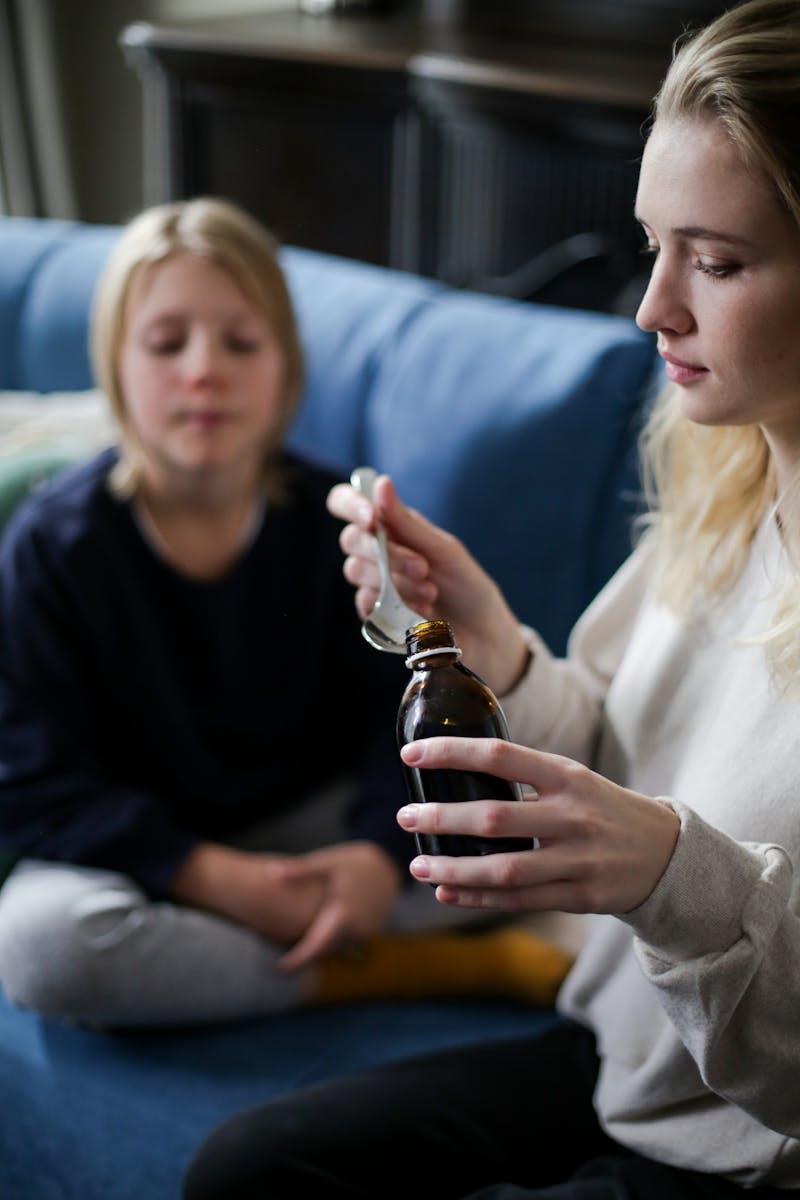 A caring mother pours medicine for her sick child sitting on a couch indoors.